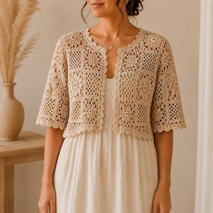 May include: A beige, open-front crochet jacket with short sleeves. The jacket features a floral and geometric pattern and a scalloped edge. The garment is worn over a cream-colored dress.