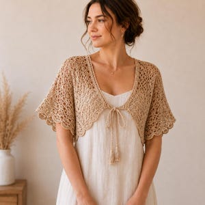Puede incluir: Un bolero de ganchillo beige de tejido abierto, con bordes festoneados y mangas cortas. El bolero se ata en la parte delantera con un cordón a juego y borlas. La prenda se lleva sobre un vestido de color claro.