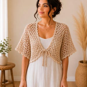 May include: A beige crochet shrug with short sleeves and a tie closure. The openwork design of the shrug is visible, and the garment is worn over a white dress. The overall style is bohemian and feminine.