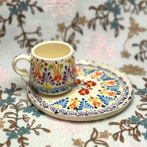 Peut inclure: Un ensemble tasse et soucoupe à café en céramique peinte à la main. La tasse et la soucoupe sont décorées de motifs floraux colorés en orange, violet, vert et bleu. La tasse a une petite anse et repose sur une soucoupe assortie.