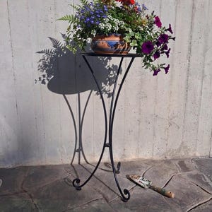 Può includere: Un supporto per piante in metallo nero con un piano rotondo che sostiene un vaso di ceramica pieno di fiori colorati. I fiori includono fiori rossi, viola, blu e bianchi. Una piccola cazzuola da giardino poggia sul terreno in pietra.