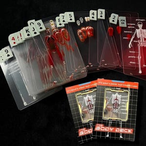 Può includere: Un mazzo di carte da gioco con carte di plastica trasparente con illustrazioni anatomiche. Le carte mostrano dettagli scheletrici e degli organi, con alcune carte che mostrano modelli anatomici rossi e bianchi. Le scatole delle carte recitano "IT'S WHAT'S INSIDE THAT COUNTS CARDS."