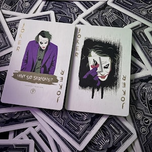 Puede incluir: Dos cartas de juego con el personaje del Joker. La carta de la izquierda muestra al personaje con traje morado y el texto Why So Serious? y Theory Eleven. La carta de la derecha muestra al personaje sosteniendo una carta pequeña con un símbolo de murciélago.