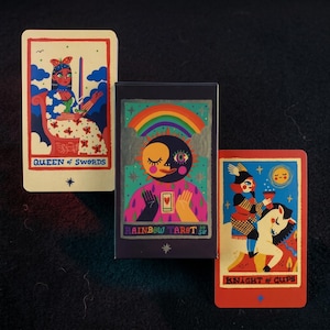 Könnte beinhalten: Ein Rainbow Tarot-Kartenspiel mit drei Karten. Die Karten zeigen farbenfrohe Illustrationen einer Königin, eines Ritters und eines Sonnen-Mond-Gesichts mit einem Regenbogen. Die Spielbox ist dunkelblau mit den Worten "Rainbow Tarot" bedruckt.
