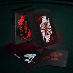 May include: A black deck of playing cards with red and white accents. The card box features a red rose design. The cards themselves have a black background with red suits and white details. Two cards are face up, displaying a rose and the word "Joker".