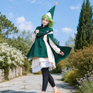 May include: A costume featuring a dark green pointed hat with a small yellow tassel, a dark green cape with black trim, a cream-colored long-sleeved dress with a white lace-trimmed hem, black leggings, and tan flat shoes with a cutout detail.