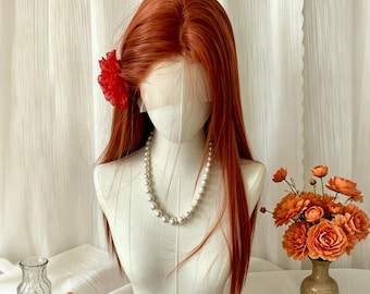 Puede incluir: Una cabeza de maniquí con cabello largo y castaño rojizo peinado con una flor roja. El maniquí lleva un collar de perlas. Flores naranjas y un pequeño jarrón están dispuestos sobre una superficie blanca frente a una cortina blanca.