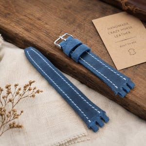 May include: A blue leather watch strap featuring white contrast stitching and a silver-toned metal buckle. A rectangular tan tag is present with the text: HANDMADE CRAZY HORSE LEATHER BUILT TO AGE, accompanied by a small leather hide icon.