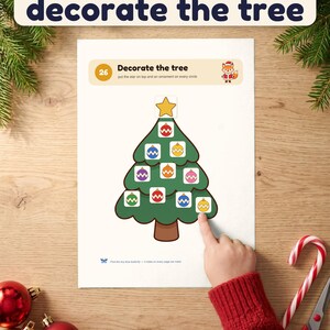 A child's hand presses an ornament onto the Decorate the tree page of the printable Christmas busy book