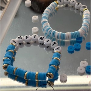 May include: Two beaded bracelets with white letter beads spelling "INFINITY" and "TSITS". The bracelets feature blue and white disc beads, accented with gold-colored spacers. The bracelets are displayed on a reflective surface.