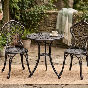 May include: A bistro set consisting of a round table and two chairs, all crafted from dark bronze-toned metal with an intricate openwork leaf and vine pattern. The table features a central support and a small ceramic cup rests on its surface.