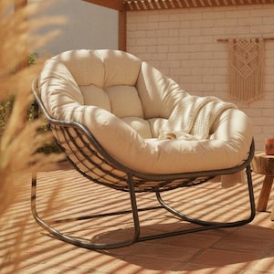 May include: A round, cream-colored tufted cushion rests inside a dark brown, woven-style metal frame chair. The chair features a curved, rocking base design and includes a soft, textured cream-colored throw blanket draped over the right side.