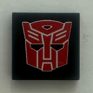 Op de afbeelding: Een vierkant, zwart embleem met een rood en wit gestileerd robotgezicht. Het robotgezicht heeft een geometrisch ontwerp met scherpe hoeken en een symmetrische indeling. Het embleem is waarschijnlijk een logo of symbool.
