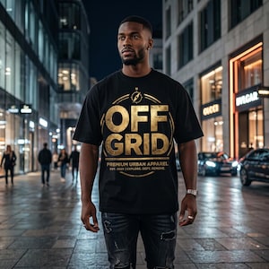 Puede incluir: Camiseta negra con un diseño dorado que dice "OFF GRID" dentro de un círculo con un diseño de brújula. El texto "PREMIUM URBAN APPAREL" y "EST. 2023 | EXPLORE, DEFY, REMOVE" están debajo. La camiseta es usada por una persona en un entorno urbano.