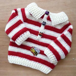 May include: A handmade crochet baby sweater with red and white horizontal stripes. The sweater has a white collar and cuffs, and is fastened with three decorative buttons. A small bee applique is sewn on the front.