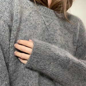 May include: A gray, oversized sweater with a crew neck and long sleeves. The knitwear appears soft and fluffy, with a subtle texture. The sweater is a neutral color, suitable for various outfits.