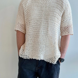Op de afbeelding: Crèmekleurig, open geweven gebreid shirt met korte mouwen. Het shirt heeft een los, luchtig ontwerp en wordt over een wit t-shirt gedragen. De persoon draagt een donkerblauwe jeans. De textuur van het shirt is zichtbaar.