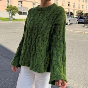 Könnte beinhalten: Ein waldgrüner Langarmpullover mit einem groben Zopfstrickmuster, rundem Halsausschnitt und lockerer Passform. Das Kleidungsstück ist aus dickem, strukturiertem Garn gefertigt und wird mit einer schlichten weißen Hose kombiniert, klassisches Design.