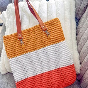 May include: A handmade crochet tote bag with a tri-color design. The top section is orange, the middle is white, and the bottom is coral. It has brown leather handles and metal clasps.