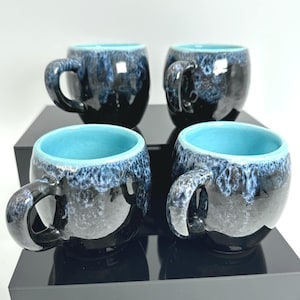 Könnte beinhalten: Ein Set aus vier abgerundeten Keramikbechern mit einer glänzend schwarzen Außenseite und einem reaktiven blauen Glasurtropfenmuster am Rand. Jeder Becher hat eine glatte, leuchtend türkisfarbene Innenseite und einen stabilen Henkel.