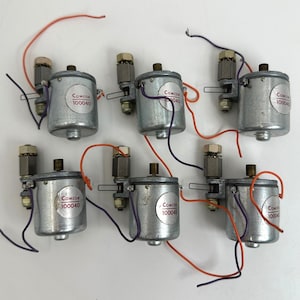 Może przedstawiać: Sześć cylindrycznych silników elektrycznych Comcor 100040 w kolorze srebrnym, ułożonych w dwóch rzędach. Każdy silnik posiada metalową obudowę, mały wałek w kolorze mosiądzu oraz pomarańczowe i fioletowe przewody z metalowymi złączami.