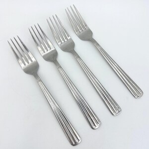 May include: A set of four stainless steel dinner forks arranged diagonally. Each fork features a polished silver finish, four tines, and a handle decorated with three vertical embossed lines that extend from the neck to the base of the handle.