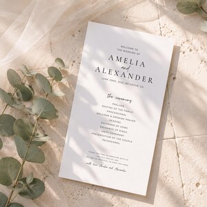 Puede incluir: Un programa de boda blanco con texto negro, con los nombres "AMELIA y ALEXANDER" y la fecha 22 de junio de 2026. El programa enumera los detalles de la ceremonia. Hojas de eucalipto y una tela transparente en el fondo.