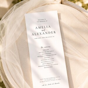 Puede incluir: Un programa de boda blanco con texto negro, con los nombres "Amelia y Alexander" y la fecha 22 de junio de 2026. El programa detalla el orden de la ceremonia, incluyendo preludio, votos y procesi&oacute;n. El programa est&aacute; colocado sobre una silla cubierta con tela transparente.