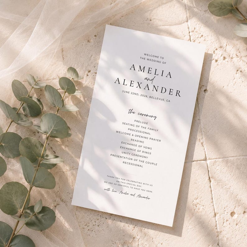 Puede incluir: Un programa de boda blanco con texto negro. El programa incluye los nombres "AMELIA y ALEXANDER", la fecha "JUNE 22ND, 2026" y los detalles de la ceremonia. Hojas de eucalipto y una tela transparente en el fondo.