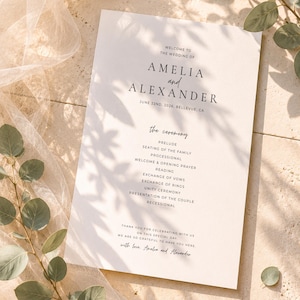 Puede incluir: Un programa de boda con los nombres Amelia y Alexander, fechado el 22 de junio de 2026. El programa detalla la ceremonia, incluyendo preludio, lecturas e intercambio de votos. El programa est&aacute; impreso en papel color crema.