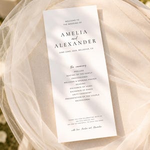 Puede incluir: Un programa de boda blanco con texto negro, con los nombres Amelia y Alexander, y la fecha 22 de junio de 2026. El programa enumera los eventos de la ceremonia, incluyendo preludio, votos y ceremonia de la unidad. El programa est&aacute; sobre una tela blanca y transparente.