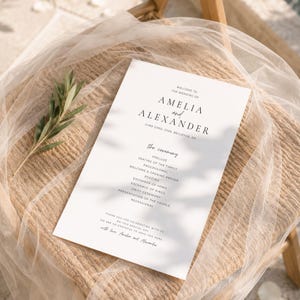 Puede incluir: Un programa de boda blanco con texto negro, con los nombres "Amelia y Alexander" y la fecha de la boda. El programa enumera el orden de la ceremonia, incluyendo preludio, lecturas y votos. Descansa sobre una silla tejida con una gasa blanca.