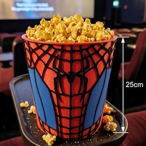 Könnte beinhalten: Ein rot-blauer Popcorn-Eimer aus Kunststoff mit schwarzem Spinnennetz-Muster und einem zentralen Spinnen-Emblem. Der Eimer ist mit Popcorn gefüllt und 25 cm hoch, was durch eine weiße Messlinie auf der rechten Seite des Behälters angezeigt wird.