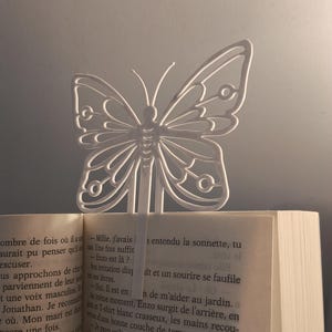 Peut inclure: Un marque-page en métal blanc en forme de papillon est inséré dans un livre. Le marque-page présente un motif ajouré détaillé. Les pages du livre sont visibles, avec du texte en langue étrangère.