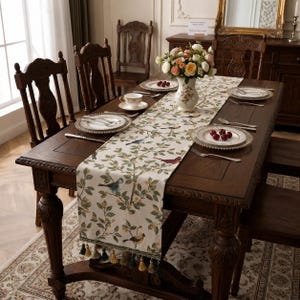 May include: A cream-colored table runner featuring a repeating pattern of colorful birds perched on leafy branches, finished with decorative green and gold tassels at the ends, laid across a dark brown wooden dining table with matching chairs.