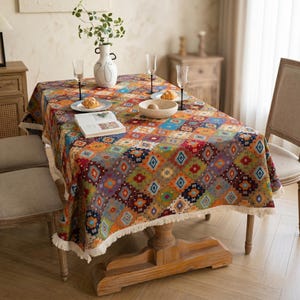 May include: A rectangular tablecloth with a colorful geometric pattern in shades of orange, blue, green, and red. The tablecloth has a fringed edge and is set on a wooden table with a vase of greenery and tableware.