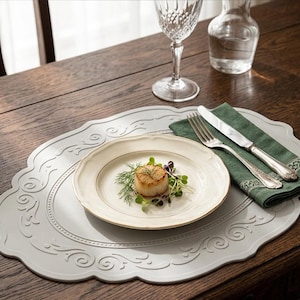 Puede incluir: Una mesa de comedor con un mantel individual gris con un diseño ornamental, un plato blanco con borde dorado y una servilleta verde con cubiertos. El plato contiene una vieira cocida y microvegetales. También hay una copa de vino y una jarra de agua.