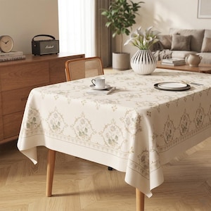 May include: A cream-colored tablecloth with a floral pattern covers a wooden table. The tablecloth has a detailed border and is adorned with a repeating design of flowers and leaves in shades of green and beige. A vase of white flowers sits on the table.