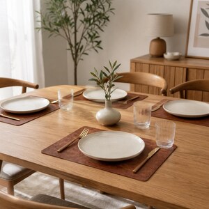May include: A light oak dining table set for four with brown leather placemats, white ceramic plates, gold-toned cutlery, and clear ribbed glass tumblers. A small white ceramic vase with a green leafy branch sits in the centre of the table.