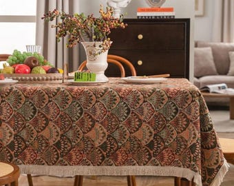 May include: A patterned tablecloth featuring a repeating leaf design in brown, green, and orange. The tablecloth has a fringed edge and is set on a wooden table. A vase of flowers and a fruit basket are on the table.
