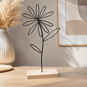 Könnte beinhalten: Eine schwarze Drahtskulptur in Form einer Blume mit einem zentralen Stiel, mehreren ovalen Blütenblättern und zwei Blättern, montiert auf einem rechteckigen, hellen Holzsockel. Die Struktur besteht aus dünnem Metalldraht mit Kontur.