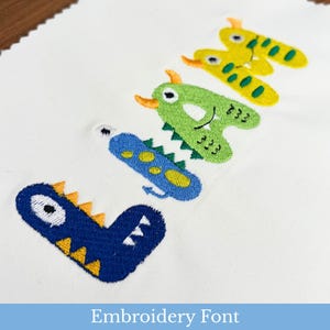 May include: A white fabric piece featuring the embroidered name Liam in a colourful monster-themed font. Each letter is designed as a unique creature with horns, teeth, and spots in shades of blue, green, and yellow, showcasing detailed stitching.