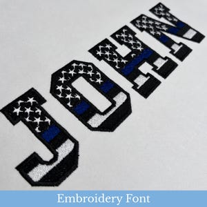 May include: The name JOHN is displayed in block letters featuring an embroidered design. Each letter incorporates a pattern of white stars on a black background, a horizontal blue stripe, and a white base, all outlined in thick black thread.