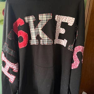 以下が含まれることがあります： 前面に「SKERS」の文字が赤、白、黒のデザインなど、さまざまな柄の生地でアップリケされた黒いパーカー。文字は赤いステッチで縁取られています。パーカーにはフードとフロントポケットが付いています。