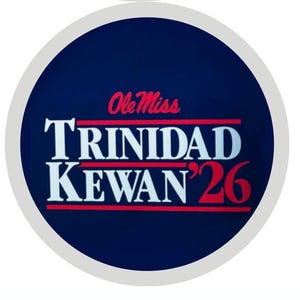 May include: A circular navy blue emblem featuring the red text Ole Miss above the white text TRINIDAD KEWAN '26. The design includes two horizontal red lines flanking the text, all enclosed within a light gray circular border on a dark background.