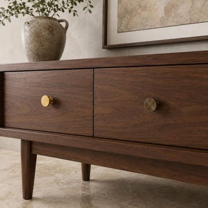 May include: A dark brown wooden console table featuring two horizontal drawers with circular brass-toned knobs. The furniture piece stands on four tapered legs and displays a visible wood grain texture across its rectangular front panels.