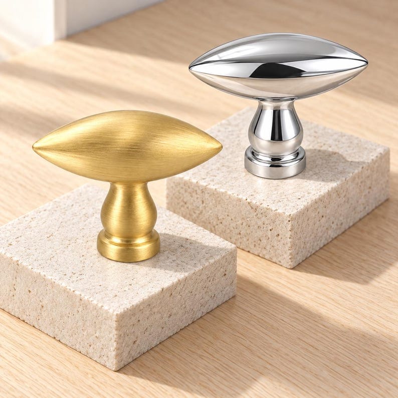 May include: Two cabinet knobs with an elongated, football-shaped top and a pedestal base. One knob features a brushed gold finish, while the other has a polished, reflective silver finish. Both items are mounted on square, speckled stone blocks.