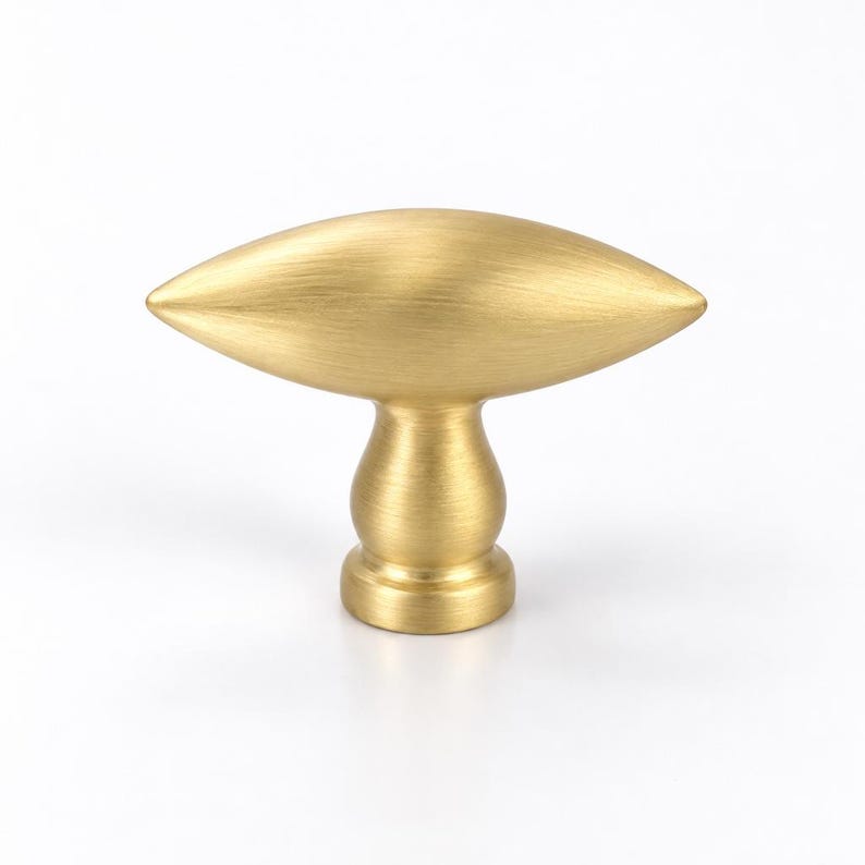 May include: A brushed brass cabinet knob featuring a horizontal, elongated, almond-shaped handle mounted on a cylindrical, tiered pedestal base. The metal surface has a soft, matte gold finish with visible fine horizontal graining throughout.