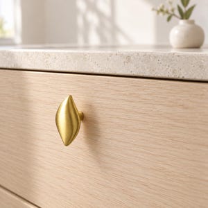 Op de afbeelding: Een geborstelde gouden kastknop met een gladde, langwerpige, bladvormige vorm bevestigd aan een lichte houten ladefront. Het metalen beslag heeft een zachte, matte afwerking en een puntige boven- en onderkant, contrasterend met de houtnerf.