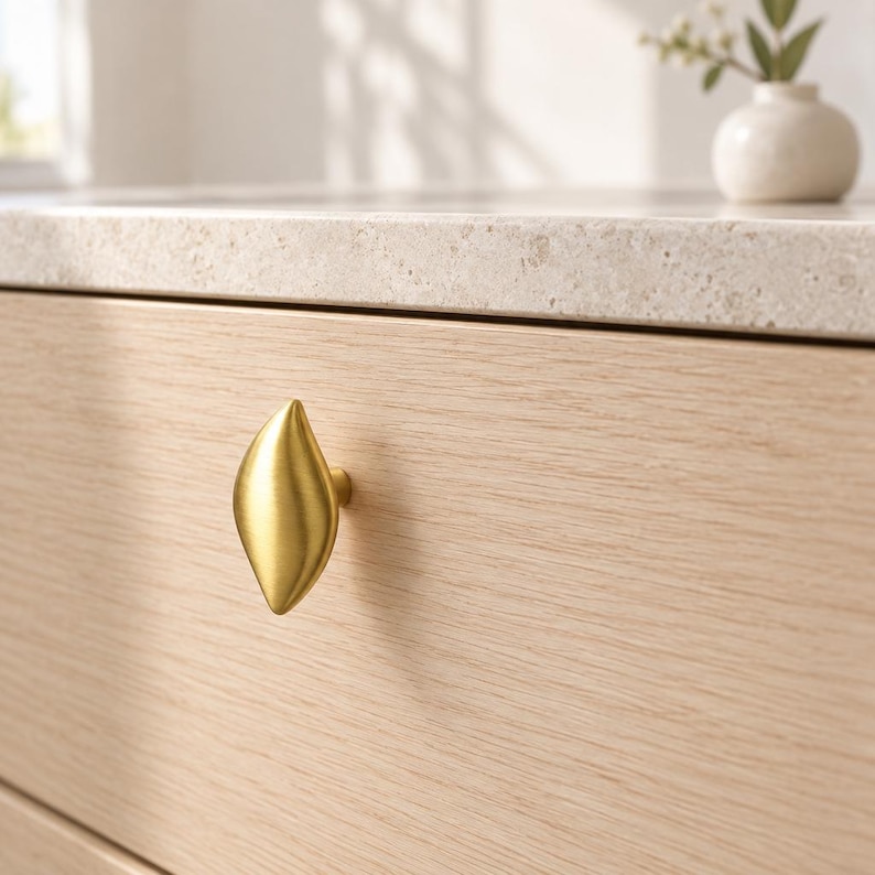 May include: A brushed gold cabinet knob with a smooth, elongated leaf-like shape attached to a light-colored wood drawer front. The metal hardware features a soft, matte finish and a pointed top and bottom, contrasting with the horizontal wood grain.